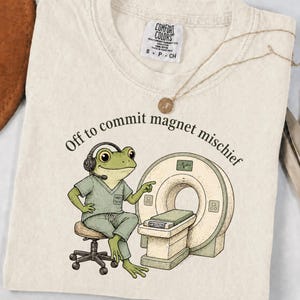 以下が含まれることがあります： クリーム色のTシャツ。ヘッドセットと医療用スクラブを着用した緑色のカエルのイラストが描かれており、MRI装置の横にあるスツールに座っています。カエルの上のテキストは「Off to commit magnet mischief」と書かれています。Comfort Colorsのラベル付き。