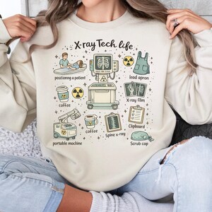 May include: Beige sweatshirt with the text "X-ray Tech life" and illustrations of x-ray equipment, coffee cups, and medical accessories. The design includes a portable machine, lead apron, and a clipboard.