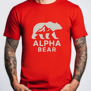 May include: A red t-shirt featuring a white graphic of a bear silhouette with mountain peaks and the text "ALPHA BEAR". The shirt is made of a soft material and has short sleeves.