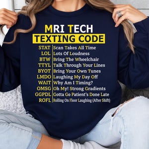 Op de afbeelding: Marineblauwe sweatshirt met de tekst "MRI TECH TEXTING CODE" in geel en wit. De sweatshirt toont een lijst met medische sms-acroniemen, waaronder "STAT" en "GGPDL".