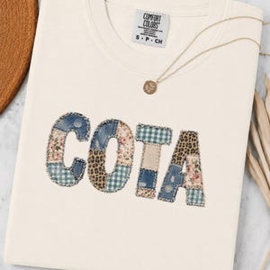 Può includere: Una t-shirt color crema con la scritta COTA in grandi lettere applicate. Ogni lettera è composta da vari ritagli di tessuto, tra cui denim blu, stampe floreali, quadretti vichy verdi e stampa leopardata, con bordi sfrangiati.