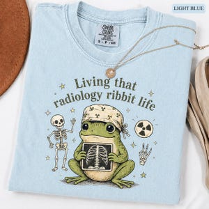 以下が含まれることがあります： 「Living that radiology ribbit life」の文字と、バンダナを巻いてレントゲン写真を持つカエルのグラフィックが描かれた、ライトブルーのTシャツ。 骸骨と放射線マークも特徴です。