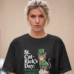May include: Black t-shirt with a white graphic of a cat wearing a green leprechaun hat and holding a pot of gold. The text on the shirt reads "St. Cat-Rick's Day: Magically Adorable."