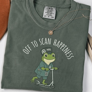以下が含まれることがあります： ダークセージグリーンのクルーネックTシャツ。スクラブとMRIキャップを身につけ、スクーターに乗ったカエルのキャラクターが描かれています。カエルの上部には「OFF TO SCAN HAPPINESS」という文字がプリントされており、Comfort Colorsのタグが付いています。