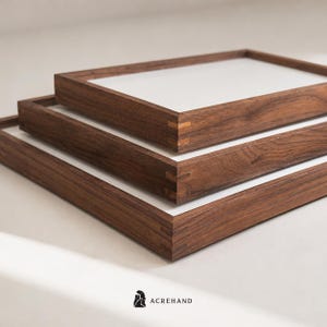 May include: A set of three rectangular wooden trays with white bases, stacked in a tiered arrangement. Each tray features dark brown wood grain frames with visible light-colored dovetail joinery at the corners. The text ACREHAND is printed below.