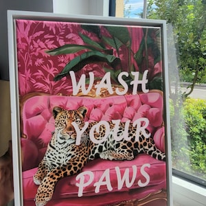 May include: A framed canvas print with a pink background and a leopard lying on a pink couch. The text "WASH YOUR PAWS" is written in white letters on the canvas.