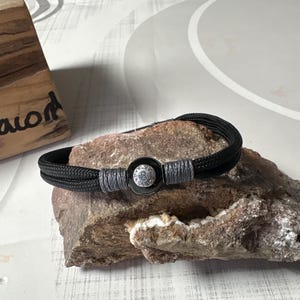 May include: Black braided bracelet with a silver-toned clasp featuring the number 8. The bracelet is accented with gray thread and is displayed on a rough, textured stone. The bracelet is a stylish accessory.