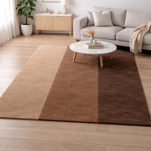 May include: A rectangular area rug with three vertical color blocks: light tan, medium brown, and dark brown. The rug is placed on a hardwood floor in a living room setting, with a sofa and coffee table.