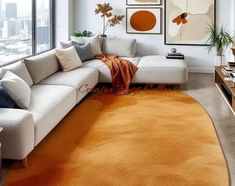 May include: An oval-shaped area rug in a solid, deep orange hue with a soft, textured surface. The rug is positioned on a light-colored floor in front of a white sectional sofa. The text Creative Rugs Collection is visible on the rug.