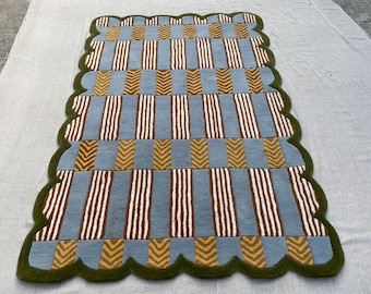 May include: A rectangular rug with a scalloped dark green border featuring a repeating pattern of light blue blocks and alternating columns of brown and white vertical stripes alongside gold chevron-patterned rectangular blocks.