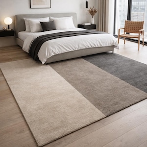 May include: A modern bedroom featuring a large bed with white bedding and a dark gray throw. A multi-tone rug in beige, brown, and gray covers the floor. A wooden chair and a window with a city view complete the scene.