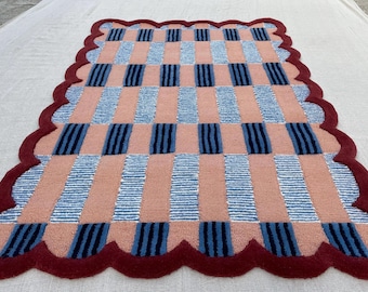 May include: A rectangular rug featuring a repeating pattern of vertical peach-colored stripes and alternating blue and white striped blocks. The rug is finished with a thick, dark red scalloped border that frames the entire perimeter.