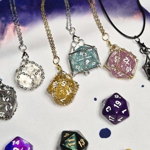 May include: A set of seven necklaces with different colored dice charms. The dice are encased in wire cages and are made of glass, glitter, and metal. The necklaces are made of silver, gold, and black chains.