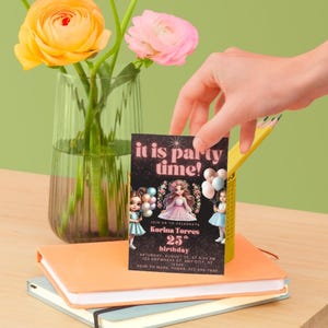 Peut inclure: Une invitation d'anniversaire avec le texte "it is party time!" présente une illustration de fée et des ballons. L'invitation est tenue par une main, avec un vase de fleurs et des cahiers en arrière-plan.