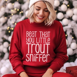 May include: A red sweatshirt with white text that reads "Beat that you little trout sniffer". The sweatshirt is worn by a person sitting in front of a Christmas tree.
