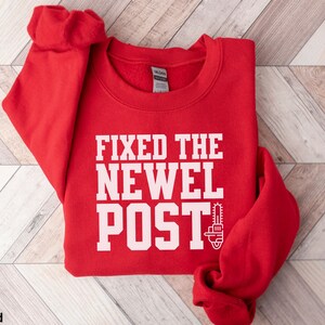 May include: Red sweatshirt with the text "FIXED THE NEWEL POST" in white with a chainsaw graphic.