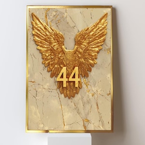 May include: A framed artwork featuring a gold-colored eagle with outstretched wings and the number "44" in gold. The eagle is set against a marble-like background with gold veining, framed in a gold border. The artwork is displayed on a white surface.