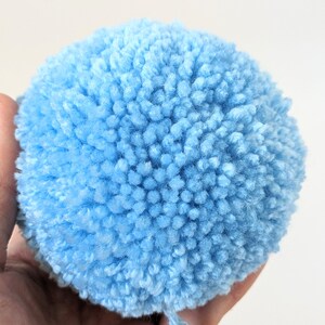 May include: A close-up of a fluffy, light blue pom-pom. The handmade craft item is round and made of soft, textured yarn. The pom-pom is held in a hand against a plain white background. The pom-pom is a craft supply.