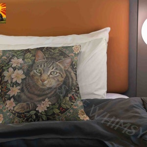 May include: A square throw pillow featuring a detailed illustration of a brown tabby cat centered within a circular frame of green leaves, small red berries, and pale pink flowers against a dark background. The cat has green eyes and whiskers.