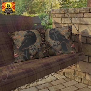 May include: Two throw pillows with a dark brown background and a black crow perched on a branch with pink flowers. The pillows are on a brown plaid swing with a wooden frame.