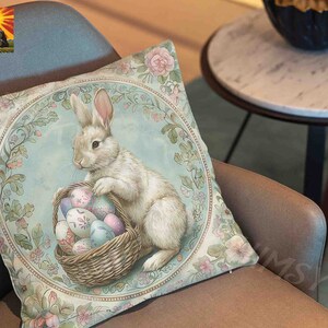May include: A decorative pillow featuring a white rabbit holding a basket of colorful Easter eggs. The pillow has a light blue background with floral designs and a gold border.