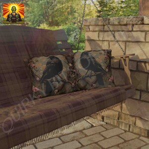 May include: Two black crow throw pillows with a floral design on a brown plaid swing. The pillows are on a wooden swing with a brown plaid cushion.