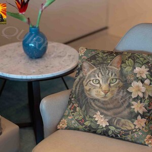 May include: A square throw pillow featuring a detailed illustration of a brown tabby cat centered within a circular frame of green leaves, small red berries, and pale pink flowers. The cat has green eyes and is looking forward.