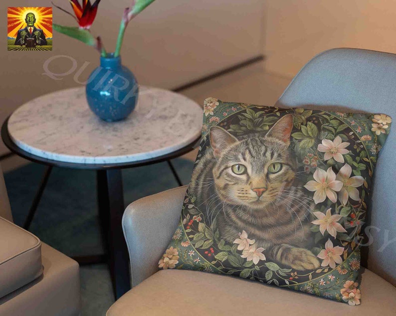 May include: A square throw pillow featuring a detailed illustration of a brown tabby cat centered within a circular frame of green leaves, small red berries, and pale pink flowers. The cat has green eyes and is looking forward.