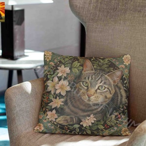 May include: A square throw pillow featuring a detailed illustration of a brown tabby cat centered within a circular frame. The cat is surrounded by light pink flowers, green leaves, and small red berries against a dark green background.