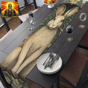 May include: A white rabbit table runner with green leaves and white flowers. The rabbit is sitting on its haunches with its ears perked up. The runner is on a dark wood table with white plates and silverware.