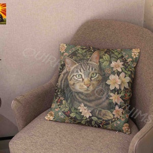 May include: A square throw pillow featuring a detailed illustration of a brown tabby cat centered within a circular frame of green foliage, small red berries, and pale pink lily flowers against a dark background on a textured fabric surface.