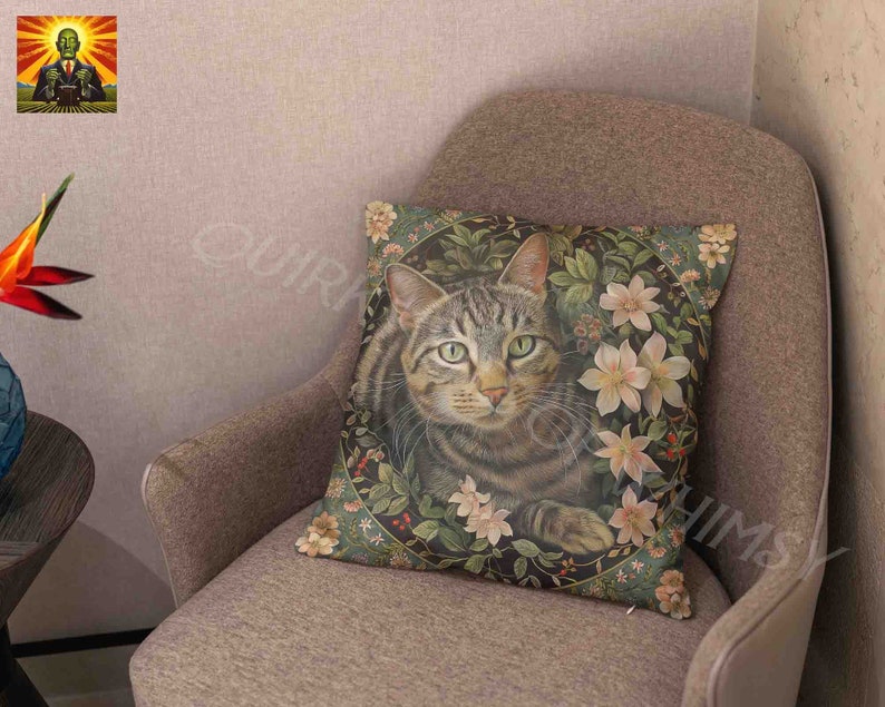 May include: A square throw pillow featuring a detailed illustration of a brown tabby cat centered within a circular frame of green foliage, small red berries, and pale pink lily flowers against a dark background on a textured fabric surface.