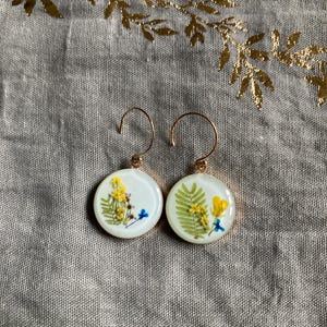 Puede incluir: Un par de pendientes redondos con ganchos dorados. Cada pendiente presenta un fondo blanco con flores amarillas y azules prensadas y hojas de helecho verdes. Los pendientes están colocados sobre una tela gris con detalles de hojas doradas.