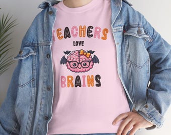 Könnte beinhalten: Rosafarbenes T-Shirt mit der Aufschrift "TEACHERS LOVE BRAINS" in Orange, Schwarz und Rosa. Die Grafik zeigt ein Gehirn mit Brille und Schleife, mit Fledermausflügeln. Die Person trägt eine Jeansjacke.