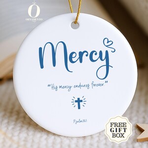 May include: A white ceramic ornament with the word "Mercy" in blue script. Below, it reads "His mercy endures forever" and features a cross symbol. A gold string is attached for hanging. Includes a "Free Gift Box" sticker.