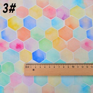 May include: A watercolor pattern of colorful hexagons on a white background. The hexagons are in shades of pink, blue, green, yellow, and orange. A ruler is placed on top of the fabric, showing the scale in centimeters.