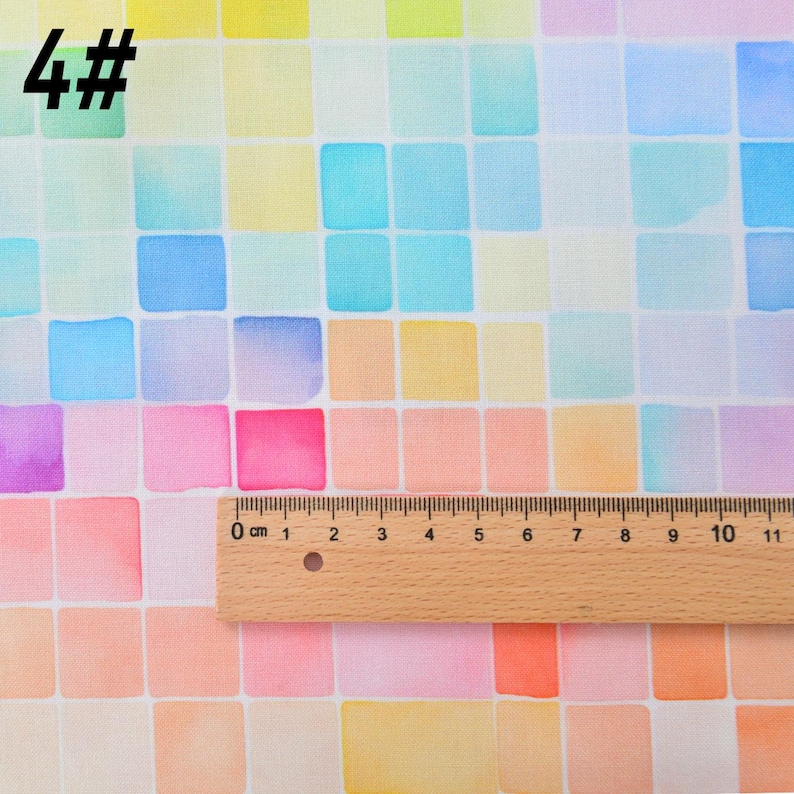 May include: A close-up of a fabric with a watercolor design of colorful squares. The fabric is white with a grid of squares in shades of blue, pink, yellow, and orange. A ruler is placed on the fabric, showing the scale in centimeters.