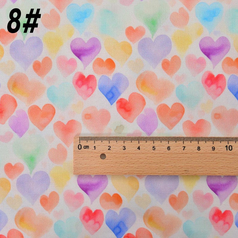 May include: A white fabric with a repeating pattern of watercolor hearts in various shades of pink, blue, yellow, and orange. The fabric is measured with a ruler in inches.