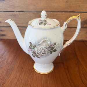 Puede incluir: Una tetera de porcelana blanca con un delicado diseño floral de rosas color crema con hojas y tallos verdes. La tetera presenta un borde dorado en el asa, el pico y la base, e incluye una tapa a juego con un pequeño motivo floral y remate.