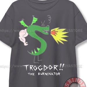 May include: A charcoal gray short-sleeve t-shirt featuring a green dragon illustration with a muscular arm, green wings, and a yellow flame coming from its mouth. Below the dragon, the text reads TROGDOR!! THE BURNINATOR in white lettering.