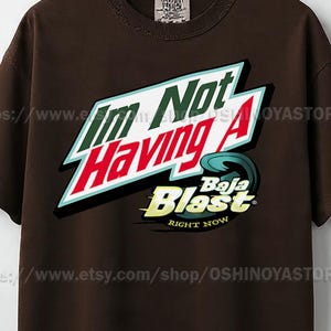 Puede incluir: Camiseta marrón con el texto "I'm Not Having A Baja Blast Right Now" en un diseño estilizado. El gráfico presenta acentos blancos, rojos, verdes y azules. La camiseta tiene cuello redondo y mangas cortas.