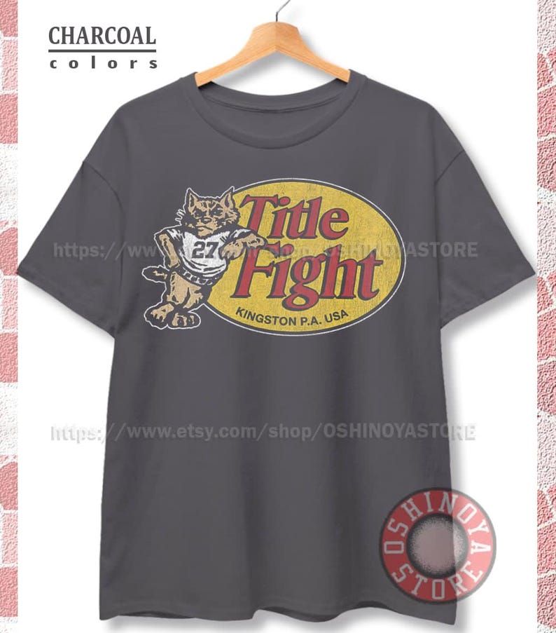 May include: A charcoal gray short-sleeve t-shirt featuring a graphic print of a cat wearing a jersey with the number 27. Next to the cat is a yellow oval containing the red text Title Fight and the words Kingston P.A. USA in black lettering.