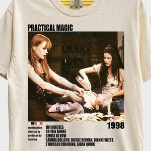 Puede incluir: Una camiseta de color crema con un estampado gráfico de la película Practical Magic de 1998. El estampado muestra a dos mujeres de pie sobre una persona tumbada en una mesa, con texto que indica el título, duración, director y reparto.