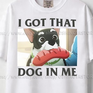 May include: White t-shirt with the text "I GOT THAT DOG IN ME" in black block letters. The shirt features a cartoon illustration of a dog holding a sausage. The dog is black and white with large eyes and a green collar.