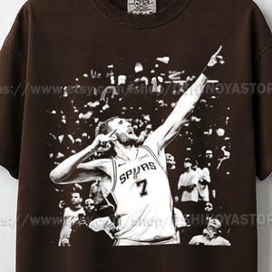 Peut inclure: T-shirt marron avec une image en noir et blanc d'un joueur de basket-ball en maillot des Spurs, numéro 7, célébrant avec le bras levé. Le t-shirt a un col rond et une petite étiquette en haut.