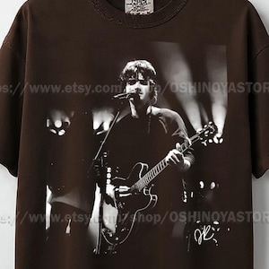 以下が含まれることがあります： ギターを弾きながら歌うミュージシャンの白黒写真がプリントされた茶色のTシャツ。画像にはミュージシャンのサインが含まれています。シャツはダメージ加工のネックラインとタグが付いています。