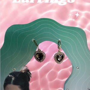 Puede incluir: Pendientes dorados en forma de corazón con una piedra central oscura y un halo de piedras más pequeñas. Los pendientes se exhiben en un soporte verde ondulado sobre un fondo rosa con el texto "Amerie's Earrings".