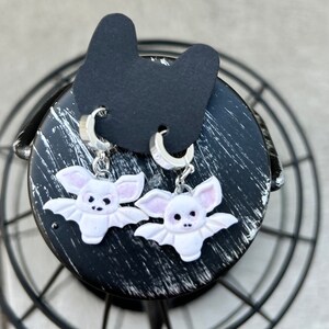 以下が含まれることがあります： ピンクのアクセントが付いた、白いコウモリ型のイヤリングのペア。 イヤリングはシルバーのフープに取り付けられており、黒い表面に表示されています。 コウモリのイヤリングは、詳細な顔と翼のデザインが特徴です。