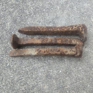Puede incluir: Tres clavos de vía oxidados y desgastados. Los clavos son de hierro y tienen una cabeza rectangular y un extremo puntiagudo. Los clavos están sobre una superficie de hormigón gris.