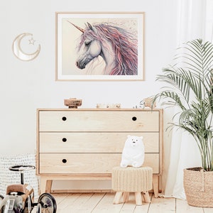 May include: A framed watercolour painting of a white unicorn with a pink and purple mane and tail. The unicorn is looking to the left and has a gold horn. The painting is on a white wall with a wooden dresser and a stuffed animal in the foreground.